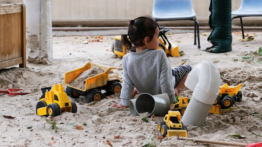 A little girl sits in a sandpit, surrounded by yellow toy dump trucks.