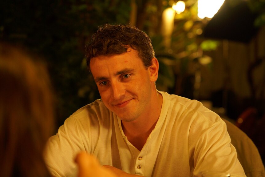 A man in his late 20s in a white shirt and smiling sitting at an outdoor restaurant at night