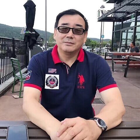 A photo of a Chinese-Australian man Yang Hengjun, sitting on an outside table, wearing a navy polo shirt with a red collar.
