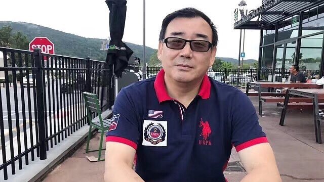 A photo of a Chinese-Australian man Yang Hengjun, sitting on an outside table, wearing a navy polo shirt with a red collar.