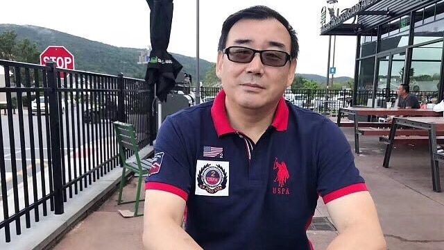 Yang Hengjun, sitting on an outside table, wearing a navy polo shirt with a red collar.