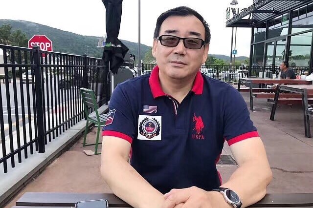 A photo of a Chinese-Australian man Yang Hengjun, sitting on an outside table, wearing a navy polo shirt with a red collar.