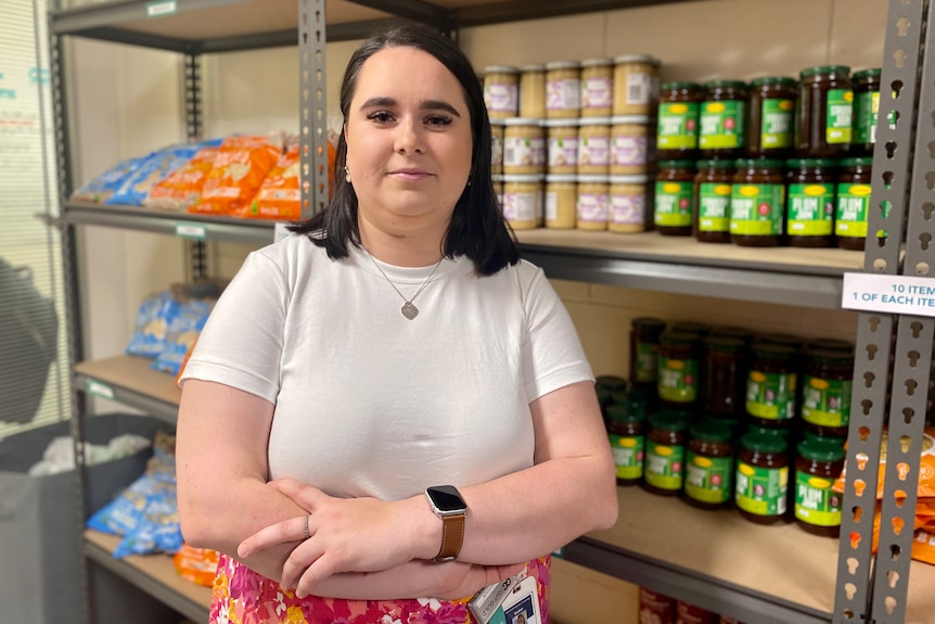 a woman in a white top in front of shelves of food jars, with her arms crossed