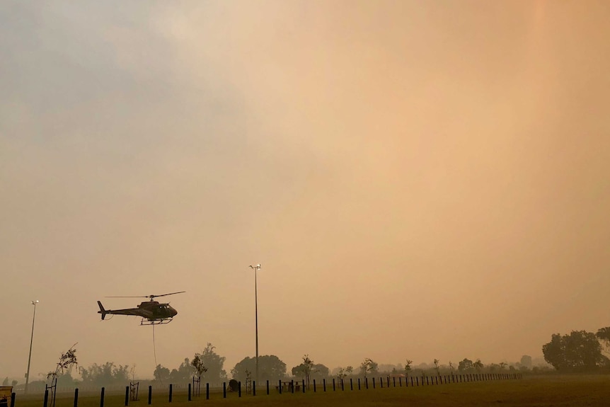 A helicopter takes off under a hazy, orange, smoke-filled sky.