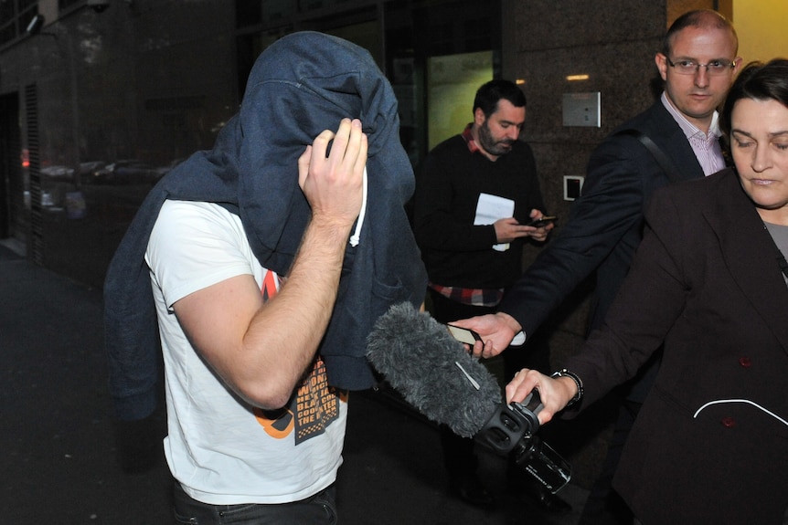 A man walks outside court with hooded jumper over his head as journalists hold up microphones towards him.