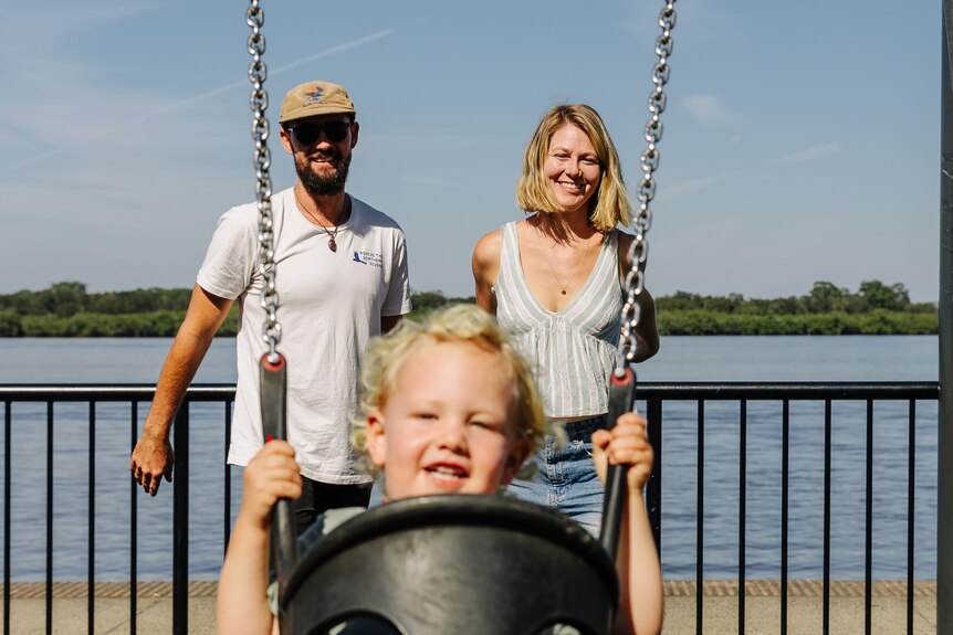 A smiling couple with their toddler on a swing in the foreground