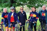 A photo of five people smiling wearing bike riding clothes with their bikes