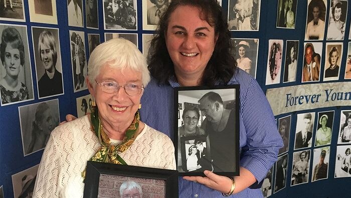 Two women stand in front of a photo exhibition showing people of all ages.
