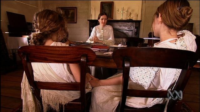 Woman in period costume talks to two girls at table