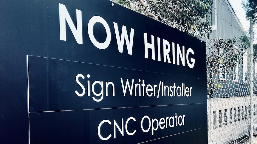 A black sign on a fence with white text that reads "Now hiring: Sign writer/installer, CNC operator, fabricator".