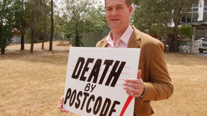 Tumut paramedic John Larter holds "Death by Postcode" sign at community protest.