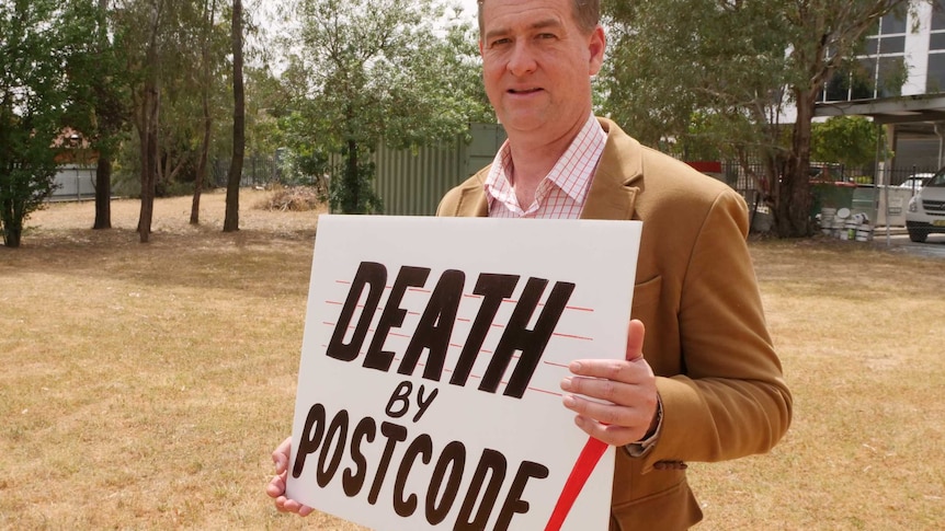 Tumut paramedic John Larter holds "Death by Postcode" sign at community protest.