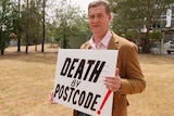 Tumut paramedic John Larter holds "Death by Postcode" sign at community protest.