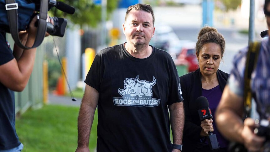 Luke Smith walks down a street wearing a black t-shirt with media walking alongside him.