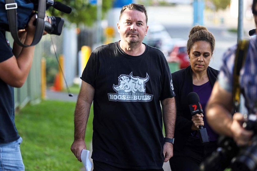 Luke Smith walks down a street wearing a black t-shirt with media walking alongside him.