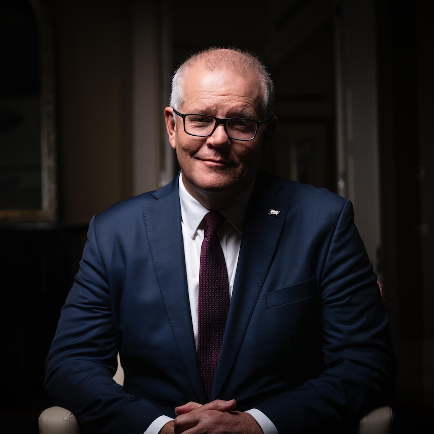 Dressed in a dark suit and tie, Scott Morrison sits in a chair, looking to camera with a slight smile.