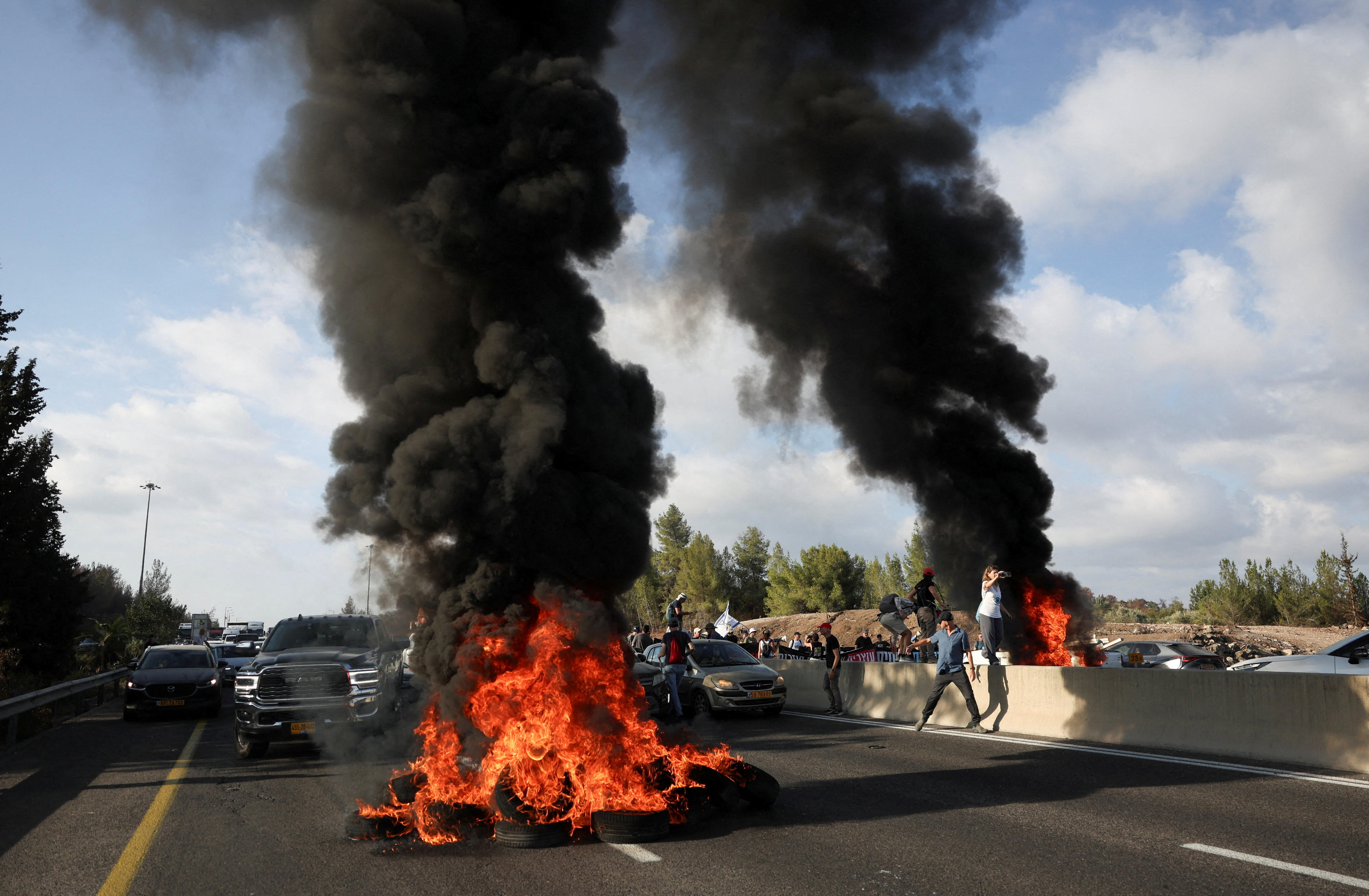 Car tyres piled up in the middle of a road and burning, with cars and people around