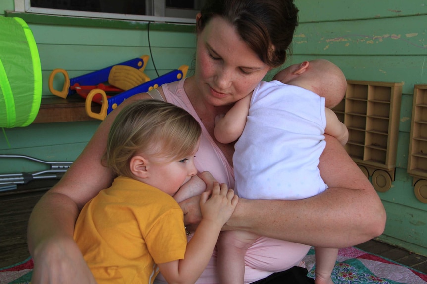 A mother breastfeeds toddler with newborn asleep on other shoulder