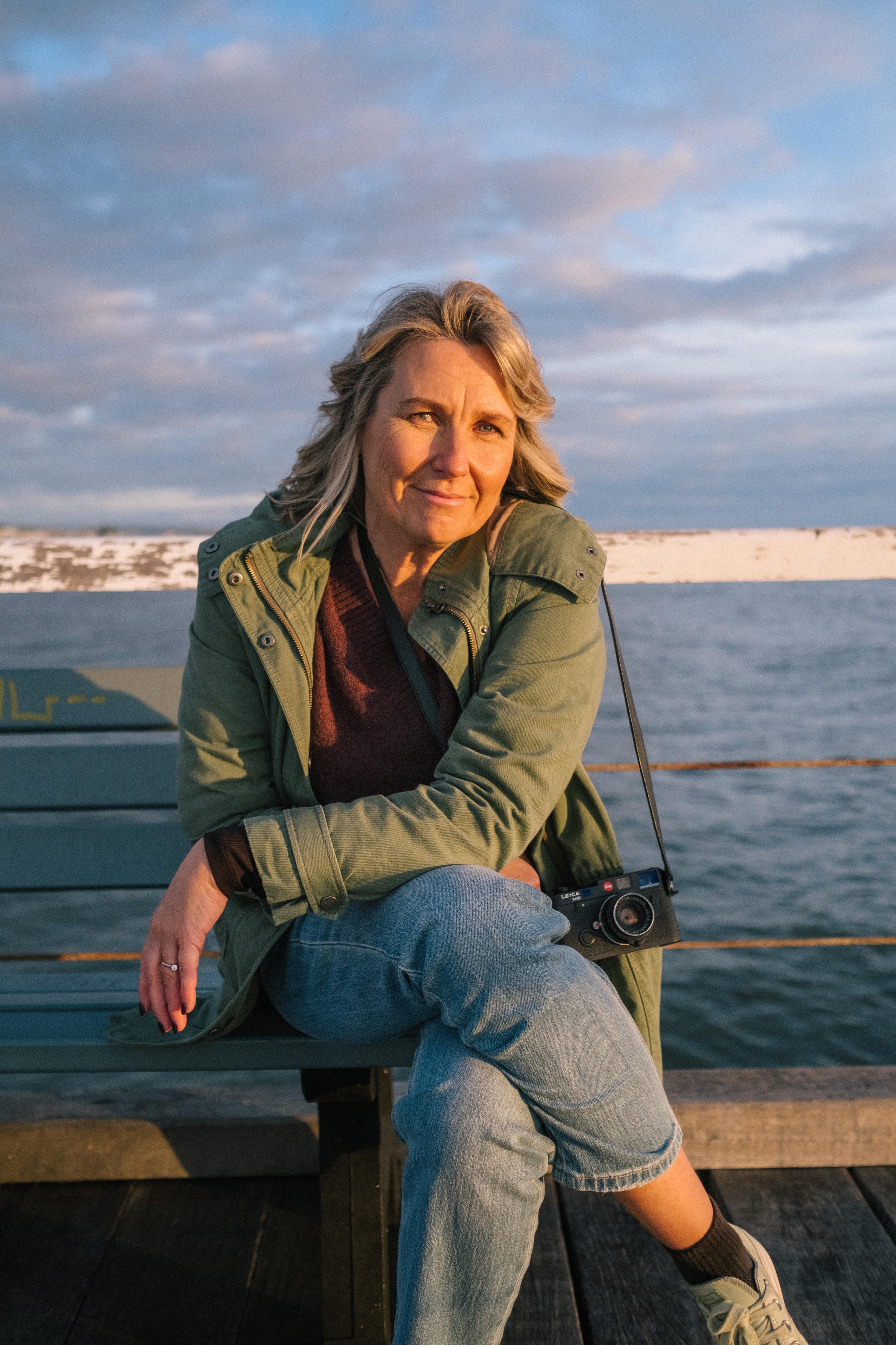 Photographer Narelle Autio sitting on a jetty at sunset, holding her camera. The light is bright on her skin and ocean dark blue