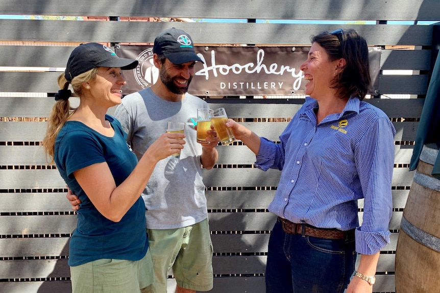 Two women and a man share a laugh as they clink their glasses of beer in a toast, in front of a sign that says Hoochery.