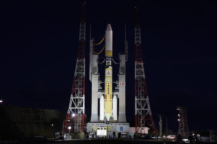 The SLIM spacecraft is mounted onto a large launcher that is illuminated while at a launch site in the dark night sky.