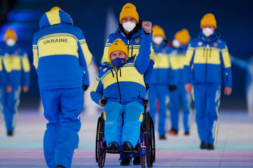 Мужчина в сине-желтом спортивном костюме в инвалидной коляске поднимает вверх кулак.