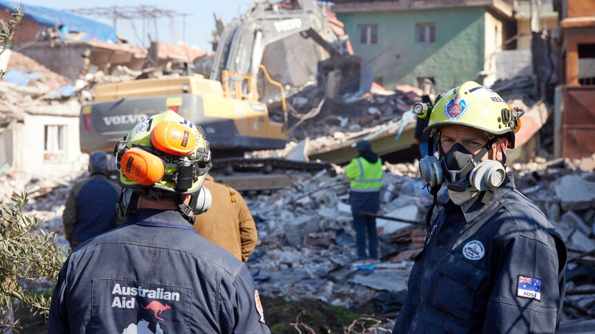 People in protective clothing with Australian logos stand in front of an excavator on building rubble.