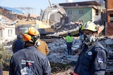People in protective clothing with Australian logos stand in front of an excavator on building rubble.