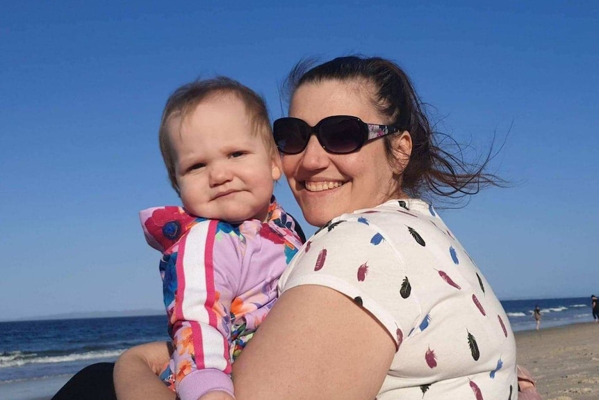 A woman in a white shirt holding her baby on the beach