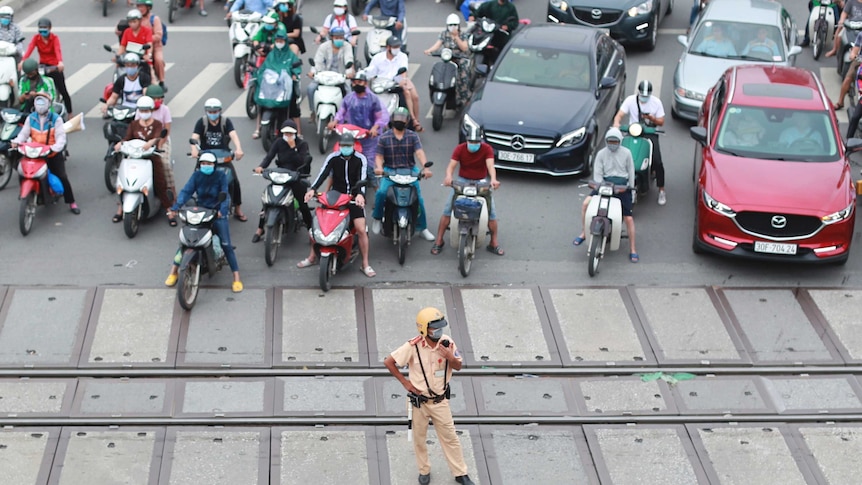 Dozens of motorbikes and cars wait at a railway crossing, managed by a policeman.