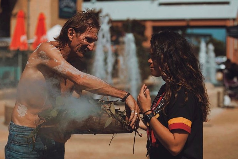 Taylah Gray stands face to face with an Aboriginal man who is holding a tray of smoking leaves between them