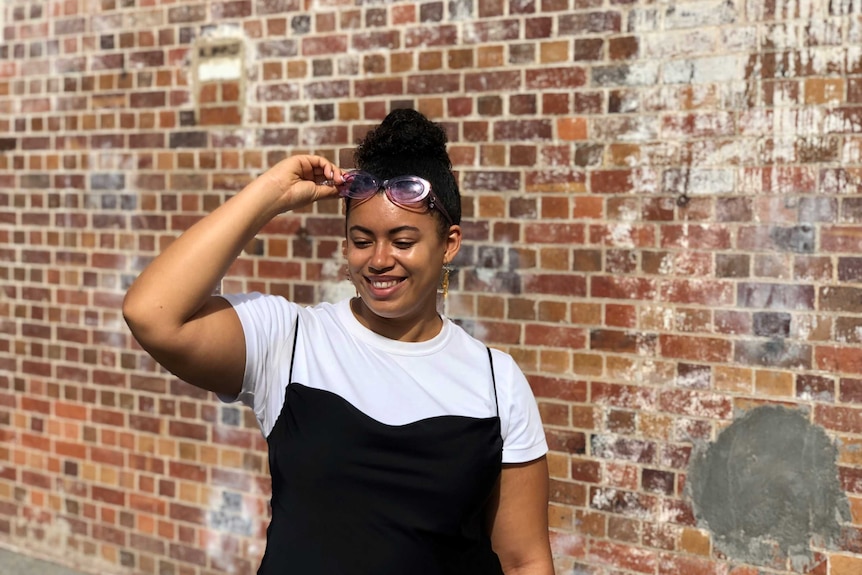 Yasmin Jeffery holds her purple glasses up as she looks away. She stands against a brick backdrop and wears a white t-shirt.