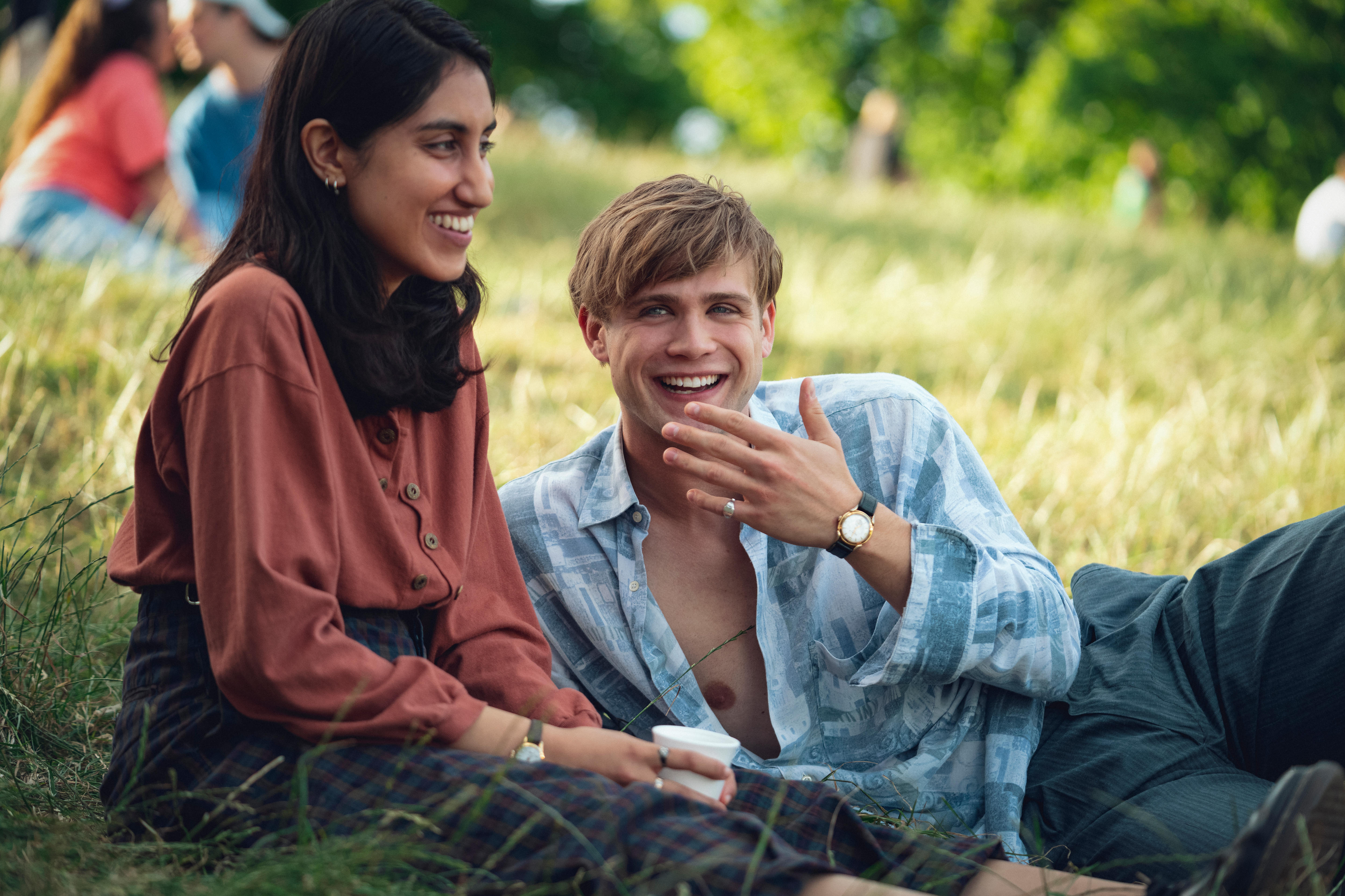 Ambika, left, sits in a grassy park with Leo laying down next to her, right. Both grin.