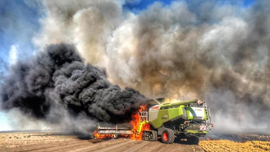 A green header is on fire in a wheat paddock. Black smoke can be seen rising into a blue sky.