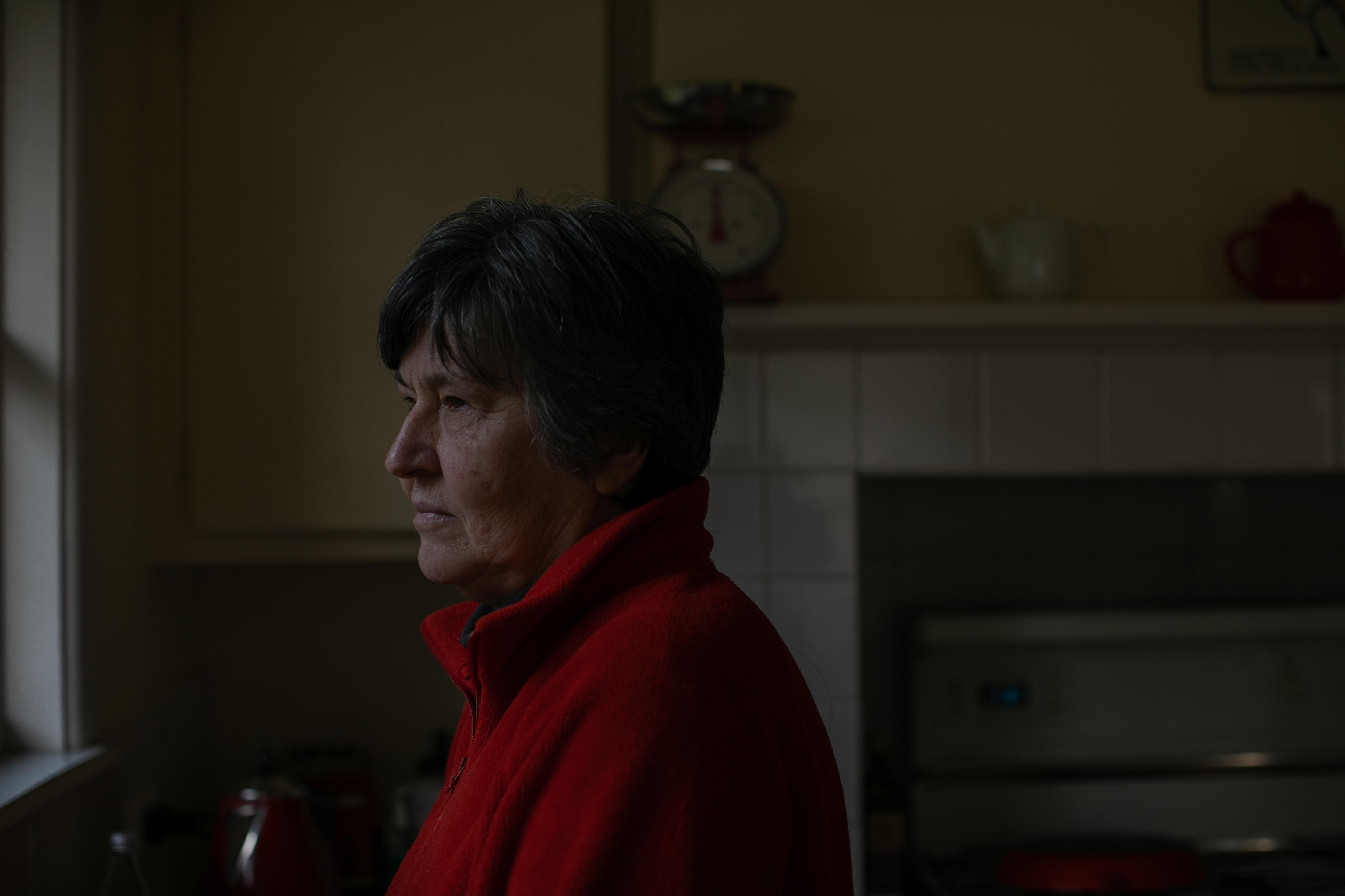 A woman wearing red polo jumper stands in a dark kitchen, looking out the window