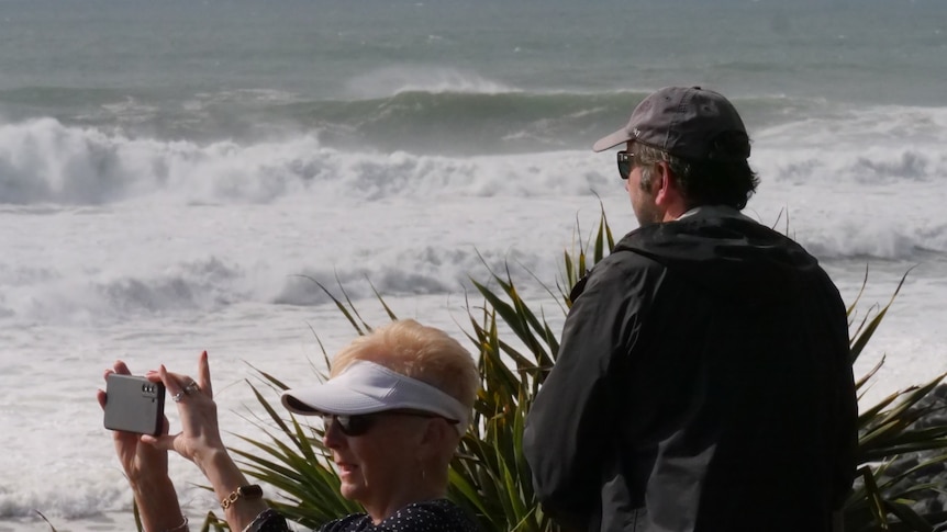 An old lady films the beach as a man looks out.