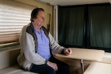 A man sits at a table inside a campervan looking downwards.