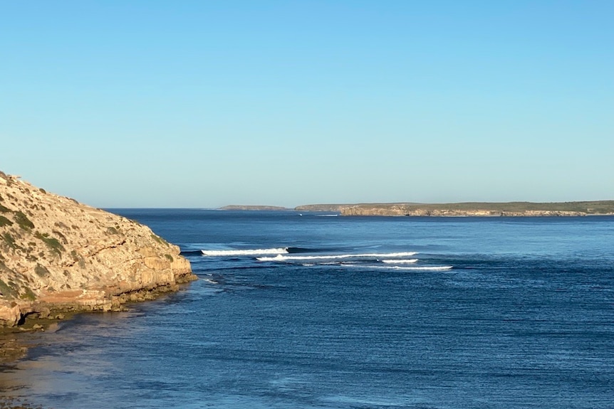 A rocky outcrop forms a break for a single white capped wave on the blue ocean. 
