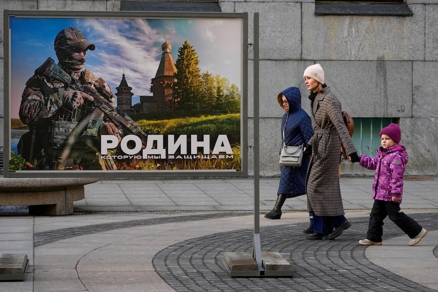 Two women a small child dressed in winter coats walk past a military sign featuring a soldier and Russian writing.