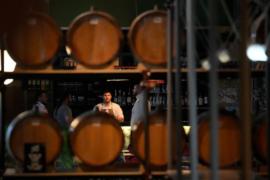 Barrels of brandy are seen on a shelf in the foreground, in front of people talking at a bar