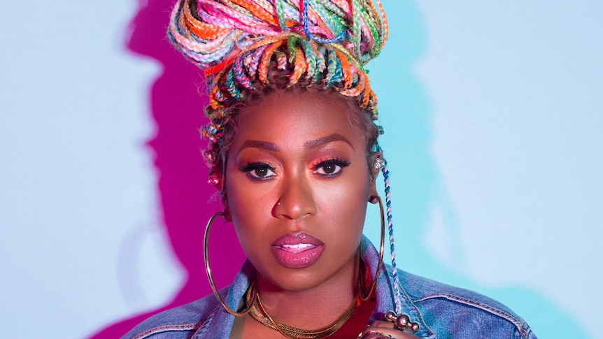 Missy Elliott with colourful braided hair wearing a denim jacket and large hoop earrings