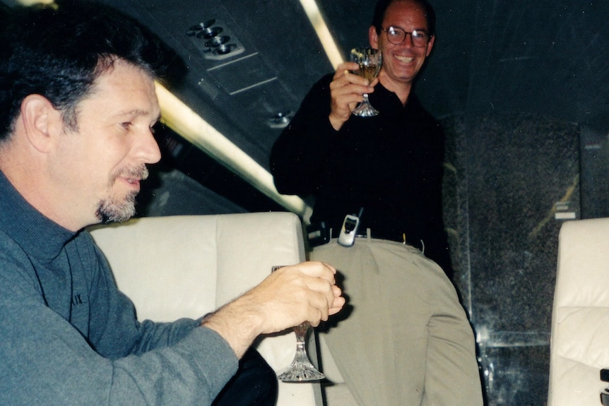 Two men wearing clothes from the early 200s smile and drink champagne.