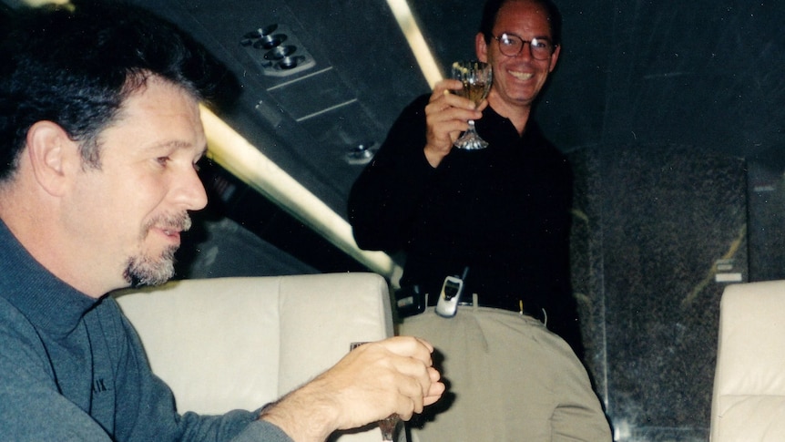 Two men wearing clothes from the early 200s smile and drink champagne.