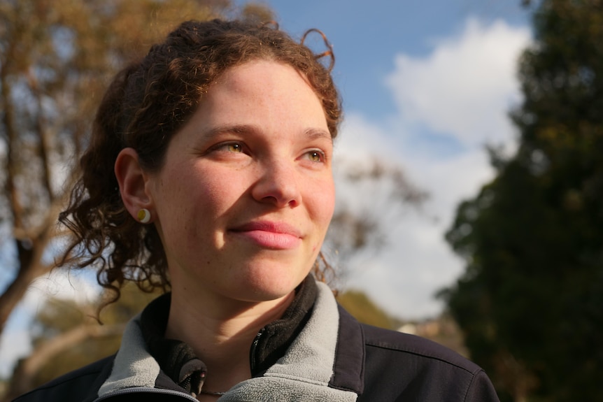 A young woman looking away from the camera with trees and sky behind her.