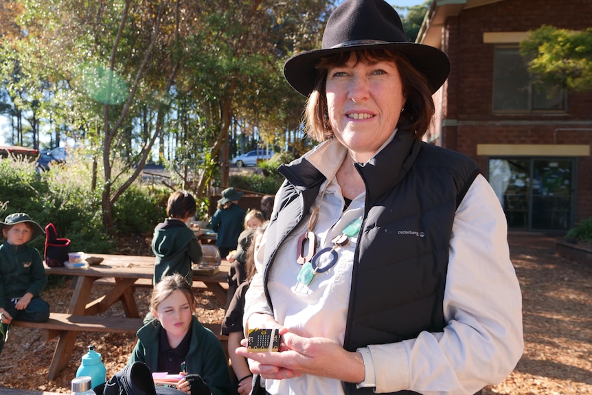 A woman stands in a school yard wearing a black hat, black vest and khaki school shirt