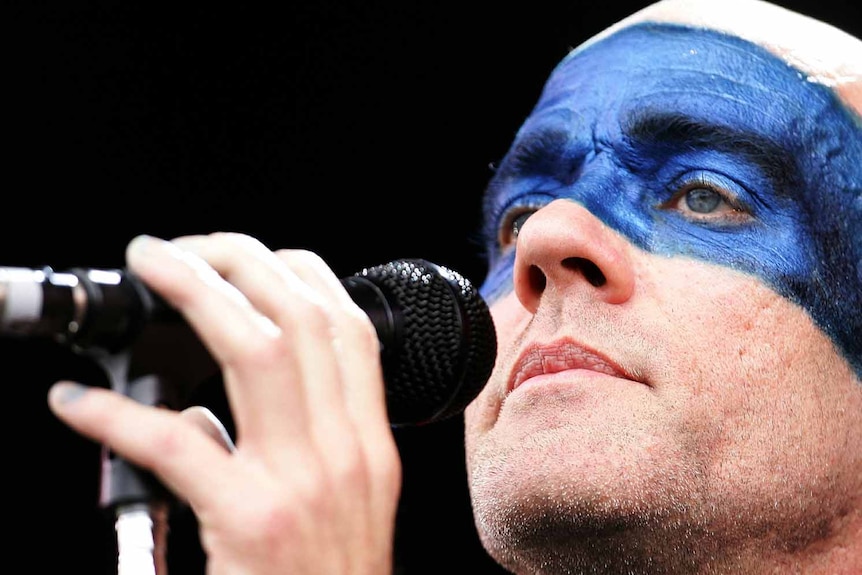 A man with blue face paint stands next to a microphone.