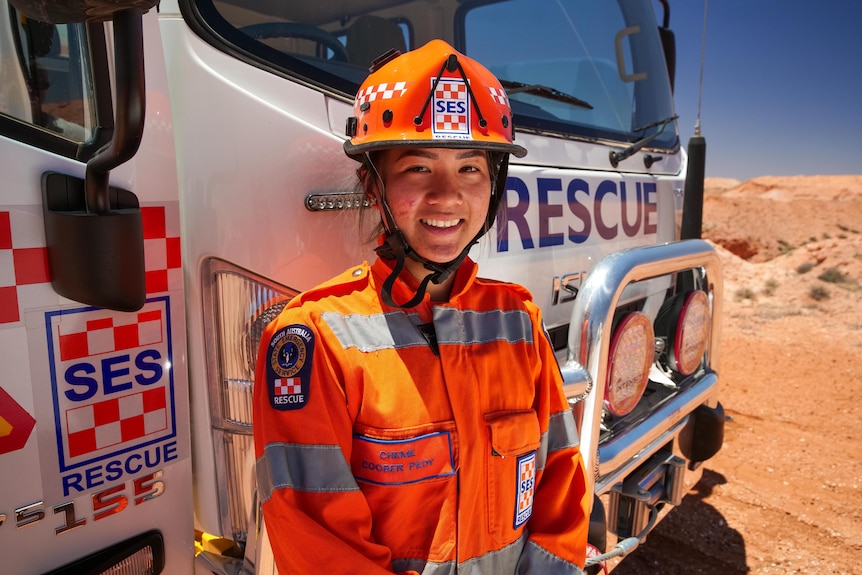 A woman smiles while wearing orange protective clothing and a helmet standing in front of a truck