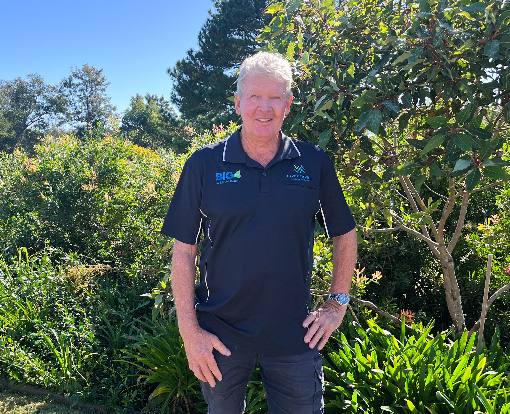 Male with grey hair and a navy t-shirt standing in front of green trees and shrubs.