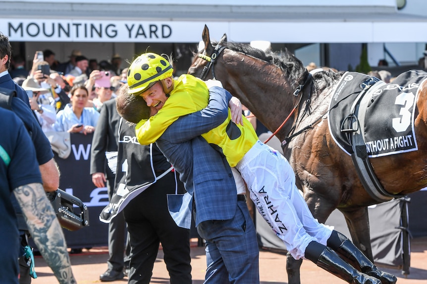 A yellow-clad jockey is hugged and lifted up by a man in the mounting yard after getting off his horse.