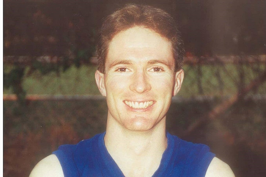 A young man in a football jumper smiles at the camera with his arms folded.